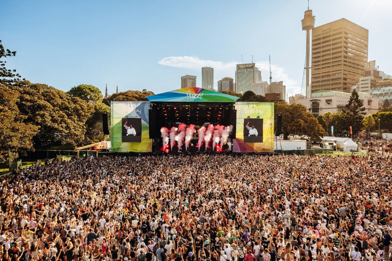 Massive crowd gathered at Sydney's Field Day music festival stage in The Domain, with pyrotechnic smoke effects and the city skyline behind.